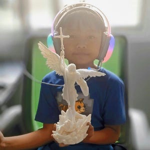 May include: A young person wearing blue headphones holds a white 3D printed angel statue with wings and a cross. The statue is standing on a base with a flower design.