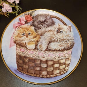 Könnte beinhalten: Ein dekorativer Keramikteller mit blauem Hintergrund zeigt eine handgemalte Illustration von drei flauschigen Kätzchen in einem Weidenkorb mit einer rosa Schleife. Die Kätzchen sind weiß, grau und orange. Der Teller hat einen goldenen Rand.