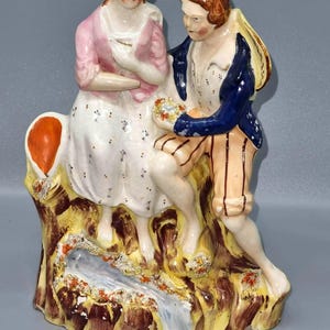 Puede incluir: Figura de porcelana vintage que representa a una pareja sentada. La mujer lleva un chal rosa y un vestido blanco, mientras que el hombre viste una chaqueta azul y pantalones a rayas. La base presenta un arroyo y follaje colorido.