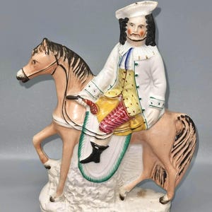 Puede incluir: Figura de cerámica de un hombre a caballo, con abrigo blanco, chaleco amarillo y sombrero tricornio. El caballo es marrón claro con marcas negras. La base dice "TOM KING". La figura tiene un escudo con un emblema negro.