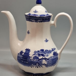 Puede incluir: Cafetera de cerámica blanca con un diseño azul de patrón Willow. La cafetera tiene un pico largo, un asa curva y una tapa con un adorno azul. El patrón azul representa un paisaje con edificios, puentes y barcos.