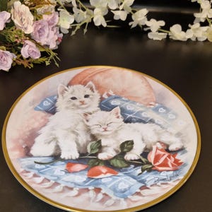 Puede incluir: Un plato de porcelana blanca con un borde dorado presenta una pintura de dos gatitos blancos durmiendo en una manta azul y blanca con una rosa roja. Los gatitos están rodeados de pétalos de rosa rosa y blanca. El plato está firmado "Brenda Walsh".