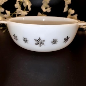 May include: White, round bowl with two handles and a snowflake pattern in black. The bowl is made of a thick, opaque material and is likely vintage. The snowflake design is repeated around the bowl's exterior.