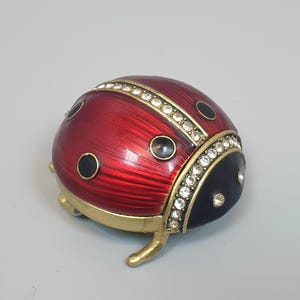 May include: A ladybird-shaped trinket box featuring a metallic red enamelled back with black spots and a black head. The piece is accented with clear rhinestones along the centre and neck, finished with a gold-toned metal base and small legs.