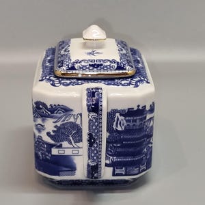 Puede incluir: Una tetera de cer&aacute;mica blanca y azul con forma cuadrada y tapa. La tetera presenta un dise&ntilde;o de paisaje azul y blanco y un ribete dorado. El asa es azul y blanca con un patr&oacute;n decorativo.