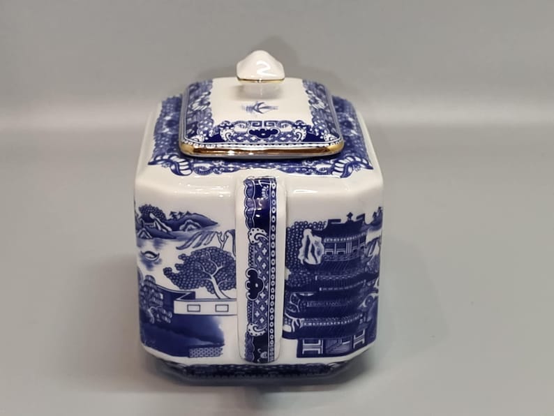 Puede incluir: Una tetera de cer&aacute;mica blanca y azul con forma cuadrada y tapa. La tetera presenta un dise&ntilde;o de paisaje azul y blanco y un ribete dorado. El asa es azul y blanca con un patr&oacute;n decorativo.