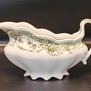 Könnte beinhalten: Weiße Keramik-Sauciere mit gewelltem Rand und gebogenem Griff. Das Boot hat ein Blumendesign in Grün- und Cremetönen. Die Sauciere hat drei dekorative Füße.