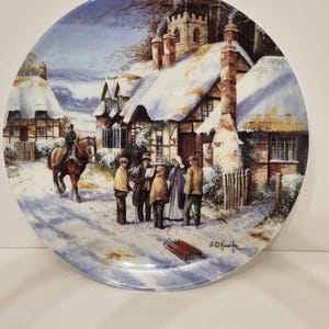 Puede incluir: Un plato navideño vintage que presenta una escena de pueblo nevado con una iglesia, casas y gente cantando villancicos. El plato está decorado con un fondo azul y blanco y un trineo rojo en primer plano.