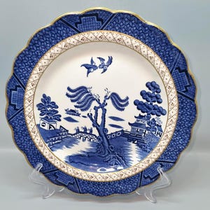 Puede incluir: Un plato decorativo azul y blanco con borde festoneado y ribete dorado. El plato presenta una escena de paisaje detallada con un puente, árboles y edificios. El borde exterior tiene un patrón geométrico.