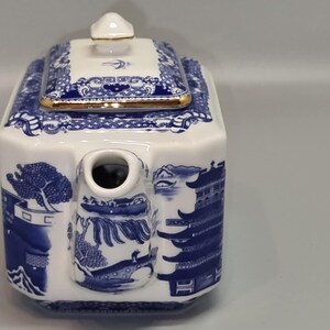 Puede incluir: Tetera de porcelana blanca y azul con forma rectangular. La tetera presenta un dise&ntilde;o de paisaje azul y blanco con un borde dorado en la tapa. El pico es cil&iacute;ndrico y la tapa tiene un peque&ntilde;o pomo.