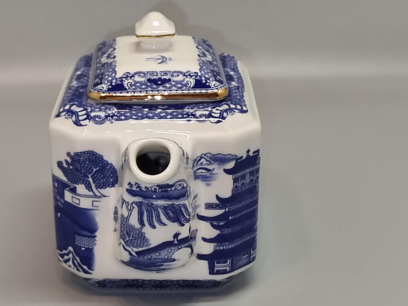 Puede incluir: Tetera de porcelana blanca y azul con forma rectangular. La tetera presenta un dise&ntilde;o de paisaje azul y blanco con un borde dorado en la tapa. El pico es cil&iacute;ndrico y la tapa tiene un peque&ntilde;o pomo.