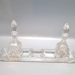 Può includere: Un set da toeletta in cristallo trasparente. Include due decanter in vetro tagliato a diamante con tappi sfaccettati, un piccolo piatto quadrato e un vassoio rettangolare. Per profumi o colonie.
