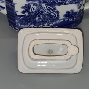 Puede incluir: Tapa de tetera de porcelana blanca, de forma rectangular con un peque&ntilde;o asa. La tapa forma parte de una tetera de porcelana azul y blanca con un dise&ntilde;o de paisaje. La tapa est&aacute; colocada sobre una superficie gris.
