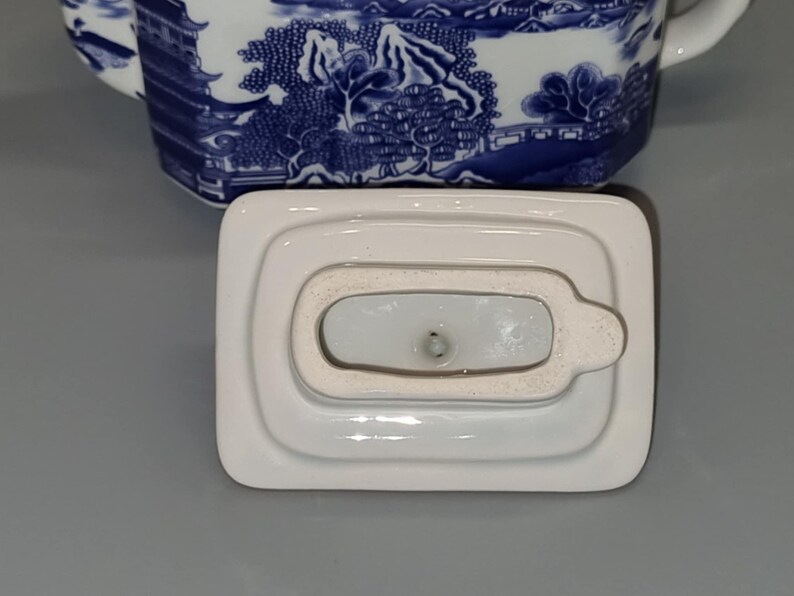 Puede incluir: Tapa de tetera de porcelana blanca, de forma rectangular con un peque&ntilde;o asa. La tapa forma parte de una tetera de porcelana azul y blanca con un dise&ntilde;o de paisaje. La tapa est&aacute; colocada sobre una superficie gris.
