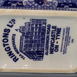 Puede incluir: Primer plano de la base de una tetera de cer&aacute;mica azul y blanca. El texto dice "RINGTONS LTD TEA MERCHANTS" y "ALGERNON ROAD NEWCASTLE ON ~ TYNE". Producida por Wade Ceramics en 1996.