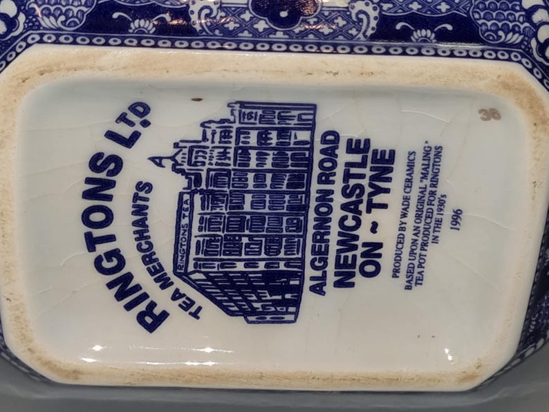 Puede incluir: Primer plano de la base de una tetera de cer&aacute;mica azul y blanca. El texto dice "RINGTONS LTD TEA MERCHANTS" y "ALGERNON ROAD NEWCASTLE ON ~ TYNE". Producida por Wade Ceramics en 1996.