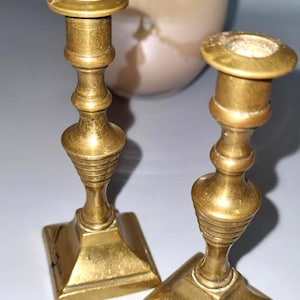 Peut inclure: Deux bougeoirs anciens en laiton avec des bases carrées et des tiges détaillées. Les bougeoirs ont un design classique et une couleur dorée chaude. L'arrière-plan présente un objet flou de couleur blanc cassé.