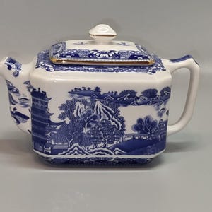 May include: A white and blue ceramic teapot with a square shape and a lid. The teapot features a blue landscape design with pagodas, trees, and mountains. The lid has a gold trim and a small white knob. The handle and spout are white.
