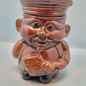 May include: Brown ceramic cookie jar shaped like a chef holding a book. The jar has a detailed face with a mustache and a chef's hat. The book reads "The Book of Biscuits How to Eat Them".
