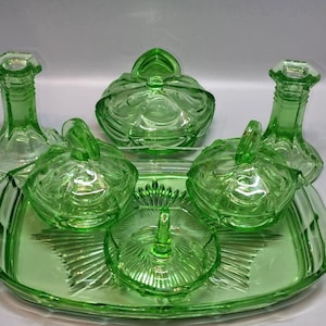 May include: A green glass dressing table set, comprising a tray, two candlesticks, and lidded dishes. The set features a ribbed design and a bright, translucent look. The tray is oval.