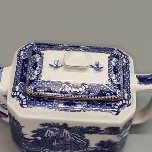 Puede incluir: Tetera de cer&aacute;mica blanca y azul con tapa rectangular. La tapa presenta un borde dorado y un dise&ntilde;o de peque&ntilde;os barcos. La tetera tiene asa y boquilla, y el cuerpo est&aacute; decorado con un paisaje. Una pieza cl&aacute;sica para los amantes del t&eacute;.