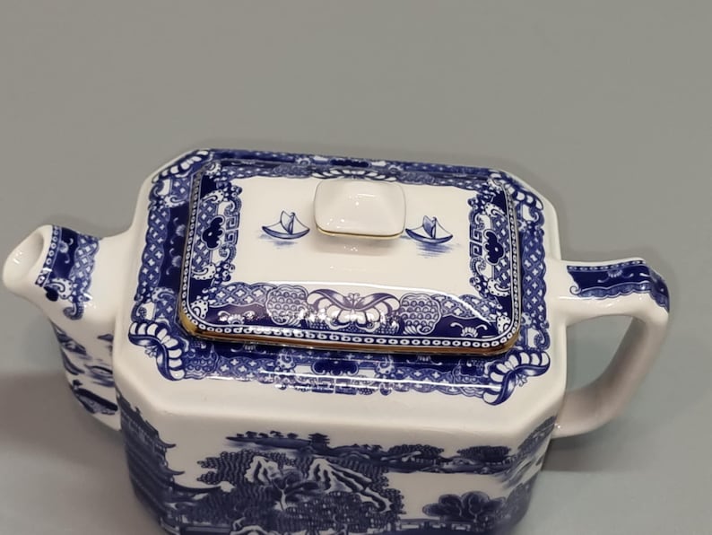 Puede incluir: Tetera de cer&aacute;mica blanca y azul con tapa rectangular. La tapa presenta un borde dorado y un dise&ntilde;o de peque&ntilde;os barcos. La tetera tiene asa y boquilla, y el cuerpo est&aacute; decorado con un paisaje. Una pieza cl&aacute;sica para los amantes del t&eacute;.