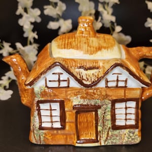 Puede incluir: Una tetera de cerámica con forma de casa de campo. La tetera presenta un tejado de paja, ventanas y una puerta, todo pintado en tonos marrones, beige y blanco. El asa y el pico también son marrones. El diseño evoca una estética rústica y campestre.