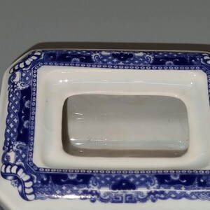 Puede incluir: Primer plano de una tapa de tetera de cer&aacute;mica blanca con una abertura rectangular. La tapa est&aacute; decorada con un patr&oacute;n floral azul y blanco en los bordes. El dise&ntilde;o recuerda a la porcelana tradicional.