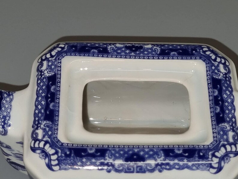 Puede incluir: Primer plano de una tapa de tetera de cer&aacute;mica blanca con una abertura rectangular. La tapa est&aacute; decorada con un patr&oacute;n floral azul y blanco en los bordes. El dise&ntilde;o recuerda a la porcelana tradicional.