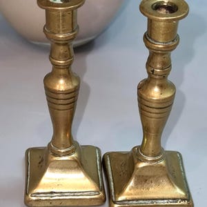 Peut inclure: Deux bougeoirs anciens en laiton. Chaque bougeoir a une base carrée, une tige cannelée et un sommet circulaire pour une bougie. Les bougeoirs sont de couleur dorée chaude, suggérant l'âge et l'utilisation.