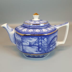 May include: A ceramic teapot featuring a blue and white transferware pattern depicting the Humber Bridge. The design includes gold trim on the lid, rim, and base. The teapot has a hexagonal body, a curved spout, a handle, and a matching lid.
