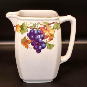 Puede incluir: Jarra de cerámica blanca con asa y boquilla. La jarra está decorada con un racimo de uvas moradas, hojas verdes y follaje naranja y amarillo. El borde tiene un ribete dorado.