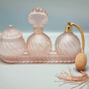 May include: A vintage pink glass vanity set comprising a perfume bottle, a lidded jar, and a decorative bottle with a seashell-shaped top. The set includes a tray and a perfume atomizer with a tassel.