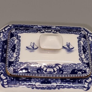 Puede incluir: Tapa de fuente de cer&aacute;mica blanca y azul con ribete dorado. La tapa presenta un dise&ntilde;o de velero azul y blanco y patrones decorativos. El asa es un peque&ntilde;o cuadrado blanco con ribete dorado.