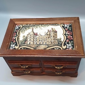 May include: A vintage wooden jewelry box with four small drawers and ornate brass handles. The top features a tapestry with a detailed castle scene, framed by a dark wood border. Floral accents surround the tapestry, adding a touch of color and elegance to the design.
