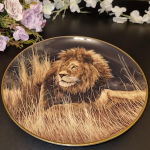 Puede incluir: Un plato de cerámica decorativo con un borde dorado presenta una pintura detallada de un león en un campo de hierba. El león mira hacia la derecha, con su melena y pelaje representados en tonos marrones y dorados.