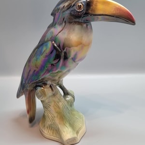 May include: A ceramic toucan ornament perched on a tree branch. The bird features iridescent plumage in shades of purple, green, and gold. Its large beak is yellow with a red tip. A decorative item.