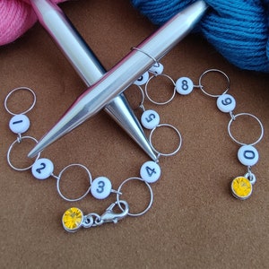 May include: Two silver knitting needles with a white and black numbered stitch marker chain. The chain has a clasp and yellow crystal beads. The numbers on the markers are 0 through 9.
