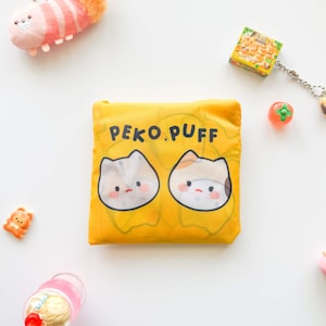 May include: Yellow tote bag with the text "PEKO.PUFF" and two cartoon cat faces. The bag is surrounded by small toys and accessories, including a plush, a miniature food box, and a small orange bear.