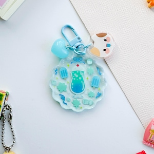 Könnte beinhalten: Ein blauer und weißer Schlüsselanhänger mit einem Anhänger eines blauen Getränks mit Eiswürfeln und einer Kirsche obenauf. Der Anhänger ist von Eiswürfeln, Sternen und Eis am Stiel umgeben. Der Schlüsselanhänger hat auch einen blauen Herzanhänger und einen weißen Anhänger mit einem braunen und weißen Hund.