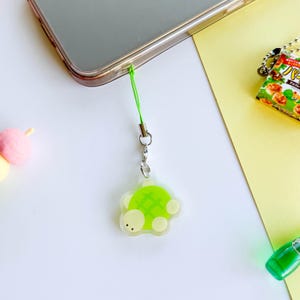 Op de afbeelding: Een groene en witte schildpadbedel op een transparante achtergrond. De bedel is bevestigd aan een groen touwtje en een zilveren ketting.