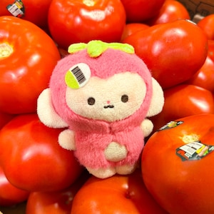 May include: A small, plush toy shaped like a monkey wearing a pink, fuzzy hood with a bright green stem on top. The toy has a cream-coloured face, dark embroidered eyes, and a small sticker with a barcode and green circle on its right side.