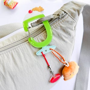 May include: A green and white cupcake-shaped carabiner clip attached to a beige bum bag. The clip has a red cherry on top and a small charm with a plush orange chick. A red cord and a light blue charm are also attached.