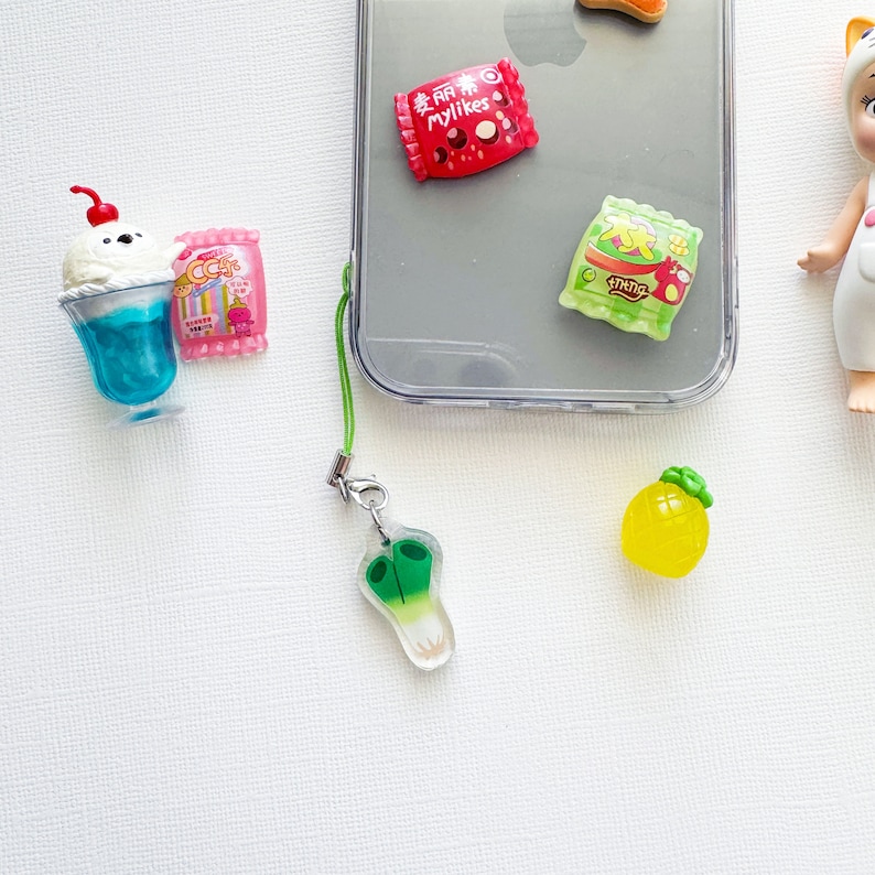 May include: A green phone charm with a white and green scallion design. The charm is attached to a green string and a silver clasp. There are other charms in the background, including a pineapple, a blue and white ice cream sundae, and a pink and white candy bar.