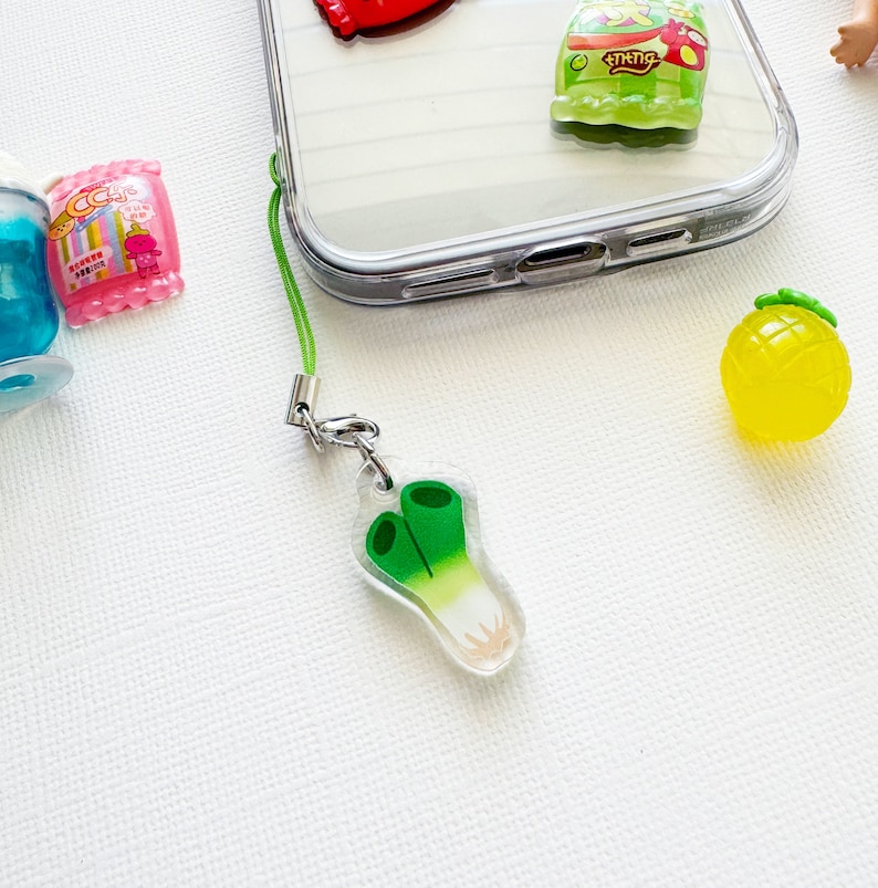 May include: A green and white leek charm attached to a green phone strap. The charm is made of clear plastic with a green leek design.