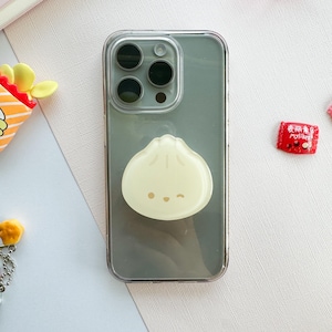 May include: Clear phone case with a light green tint, featuring a dumpling-shaped phone grip. The grip is a pale yellow color with a cute, winking face. Other small, colorful accessories are visible in the background.