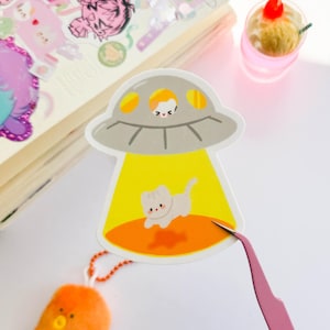 May include: A sticker featuring a cartoon UFO with a white cat inside and another cat in the beam of light. The UFO is gray with yellow accents, and the beam is yellow. The sticker is on a white surface.
