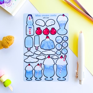 Puede incluir: Una hoja de pegatinas con ilustraciones de refresco de crema azul. Los diseños incluyen una botella de refresco, helado, paletas, nubes y flotadores de refresco. El texto "blue cream soda" y "peko. puff" son visibles. Un bolígrafo blanco y pinzas están al lado.
