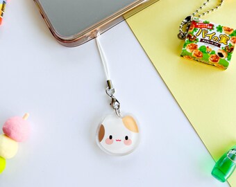 May include: A white and brown cat charm with a pink blush on its cheeks, attached to a white phone strap. The charm is made of clear plastic and has a cute cartoon style.