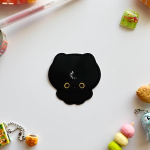 Può includere: Un adesivo nero a forma di gatto con occhi gialli e un simbolo di caricamento. L'adesivo è circondato da altri piccoli oggetti, tra cui una penna, nastro adesivo e snack, il tutto su una superficie bianca.